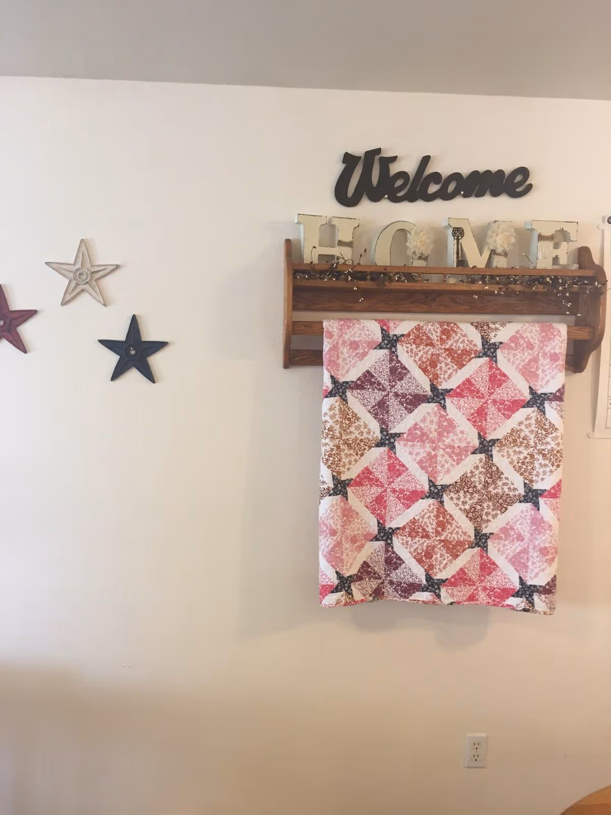 A wall decorated with three star-shaped ornaments in red, beige, and navy blue on the left side. In the center, a wooden rack holds decorative letters spelling 'HOME' with a 'Welcome' sign above it. A colorful quilt with a pattern of pink, purple, and brown squares hangs from the rack.