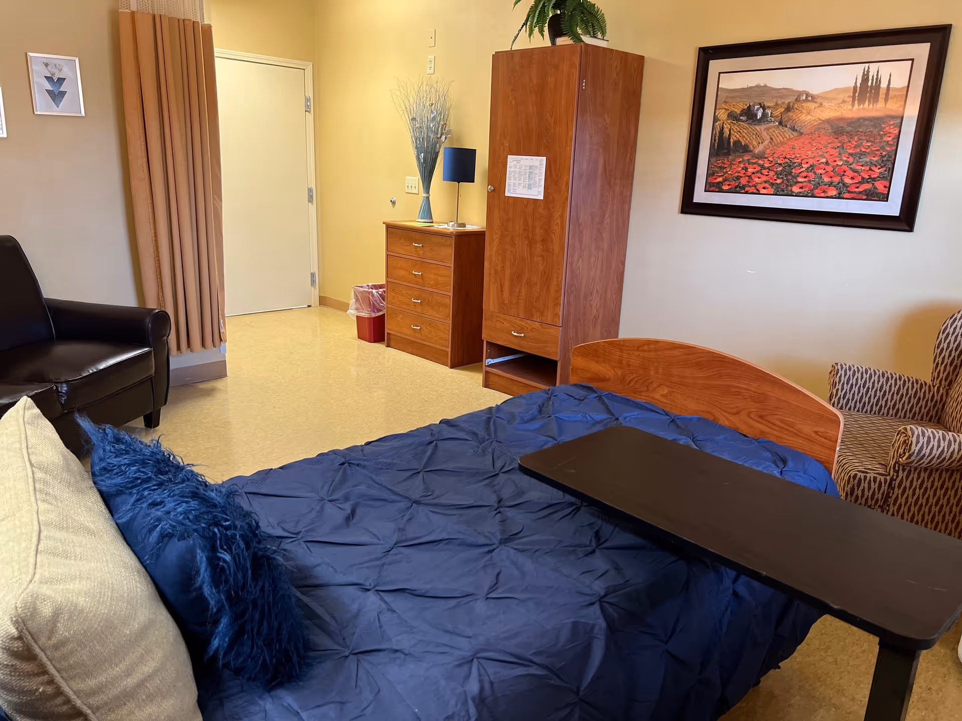 A senior living facility bedroom with a bed covered in a blue comforter and pillows, a black overbed table, a brown armchair, a black leather chair, a wooden dresser with a lamp and decorative vase, a wooden wardrobe, and a framed landscape painting on the wall.