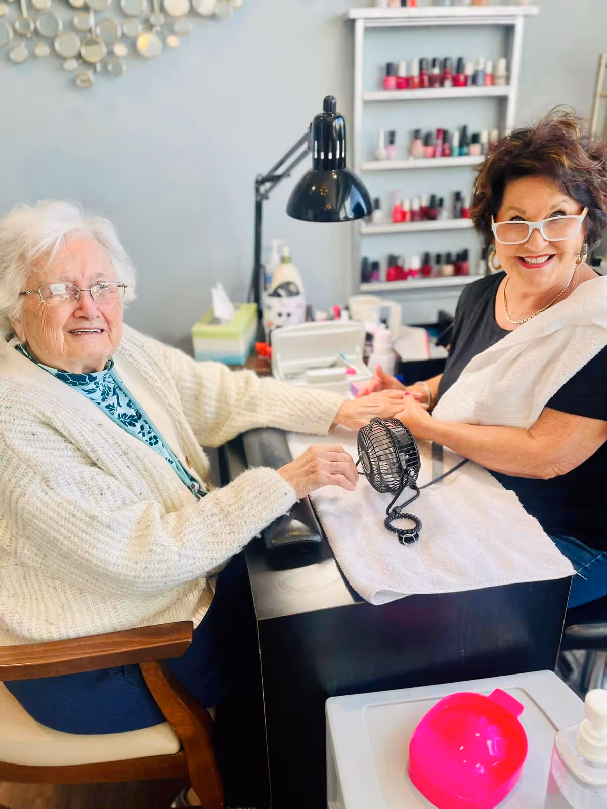 An elderly woman with white hair and glasses is sitting at a manicure station, smiling while a nail technician with short dark hair and white glasses is holding her hand and smiling. The background shows shelves with various nail polish bottles and manicure supplies on the table.