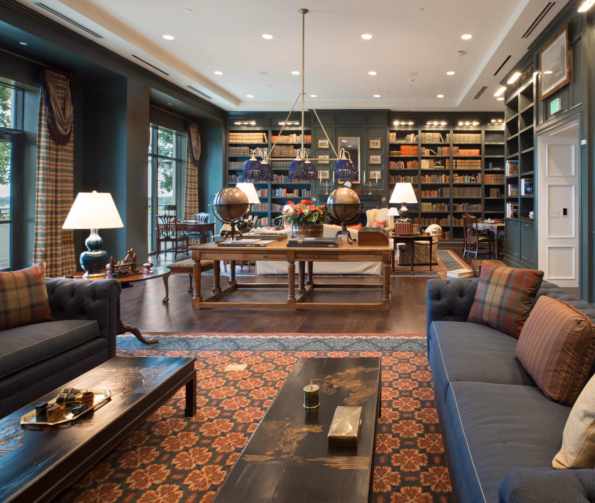 A spacious and elegant living room with dark wood floors, large windows with plaid curtains, and built-in bookshelves filled with books. The room features two dark blue sofas with plaid and striped pillows, two wooden coffee tables, a large wooden table with two globes and a flower arrangement, and several table lamps providing warm lighting.