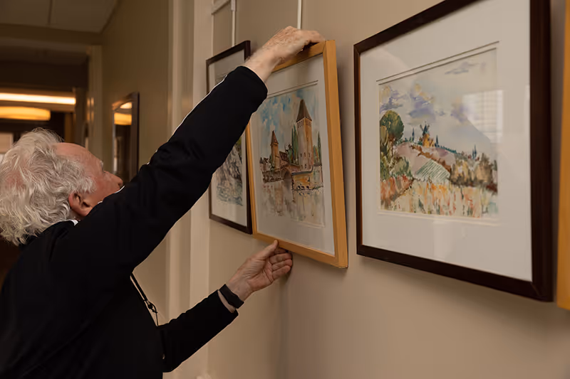 An elderly person with white hair is hanging a framed watercolor painting on a beige wall in a hallway. The painting depicts a scenic view with buildings and water. Other framed paintings with similar watercolor landscapes are already hung on the wall.