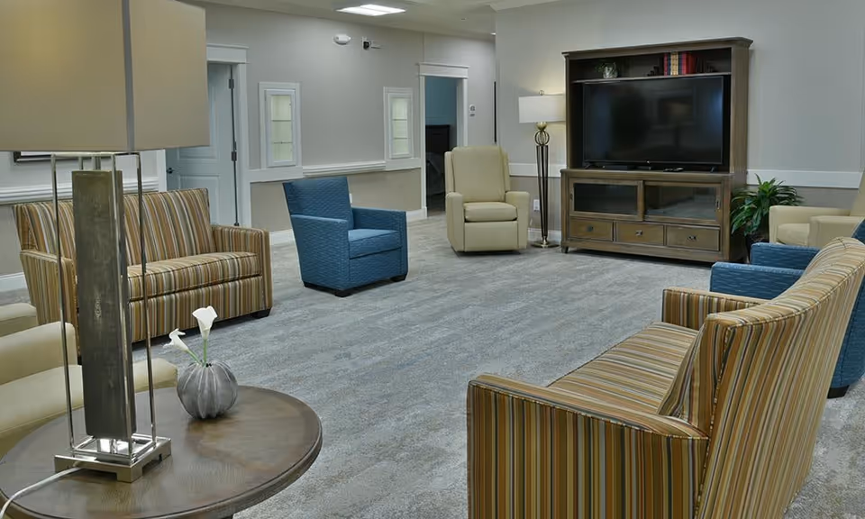 Spacious senior living common room with striped and upholstered chairs arranged around a TV cabinet and lamps.