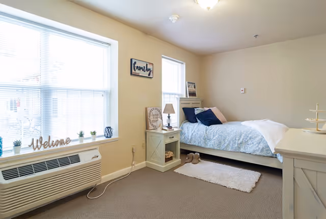 Bright furnished bedroom with a bed, nightstand, windows with blinds and decorative accents.