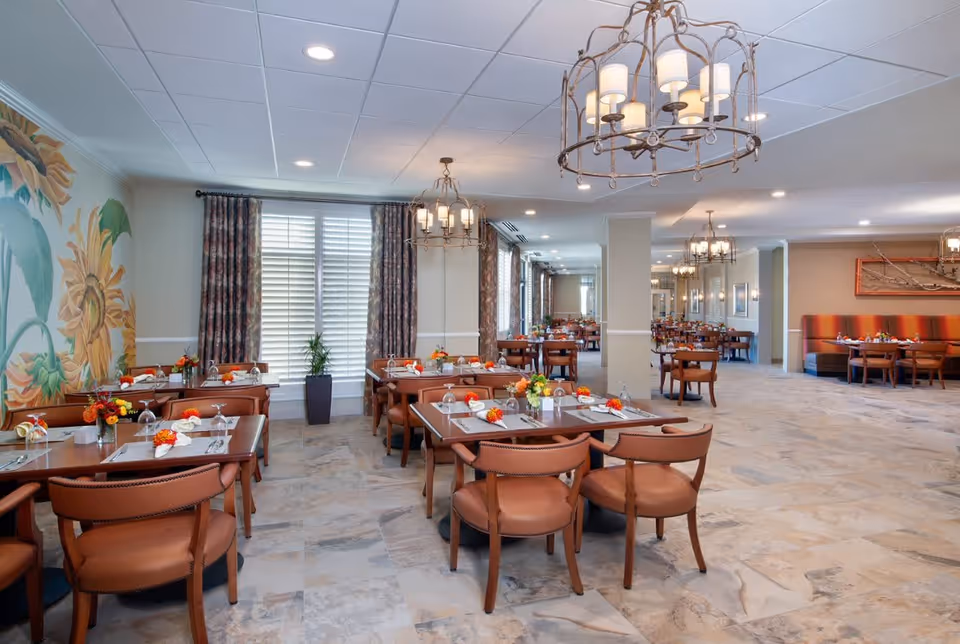 A spacious dining room with multiple wooden tables and brown leather chairs. Each table is set with placemats, napkins, glasses, and small floral centerpieces. The room features large windows with patterned curtains, decorative chandeliers hanging from the ceiling, and a mural of sunflowers on one wall. The floor is tiled with a light stone pattern.