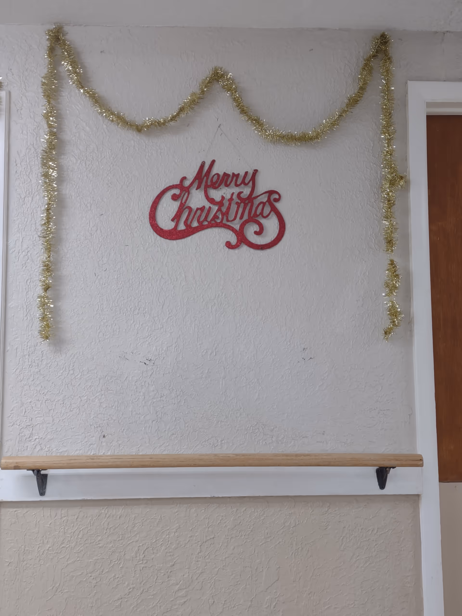 A textured interior wall decorated with a red 'Merry Christmas' sign and draped gold tinsel above a wooden handrail.