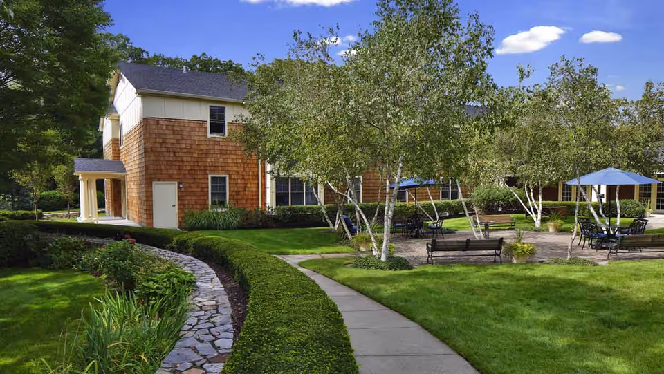 Outdoor garden area at a senior living facility with green grass, trees, benches, and tables with blue umbrellas. A two-story building with brown siding and white trim is visible in the background under a blue sky with a few clouds.