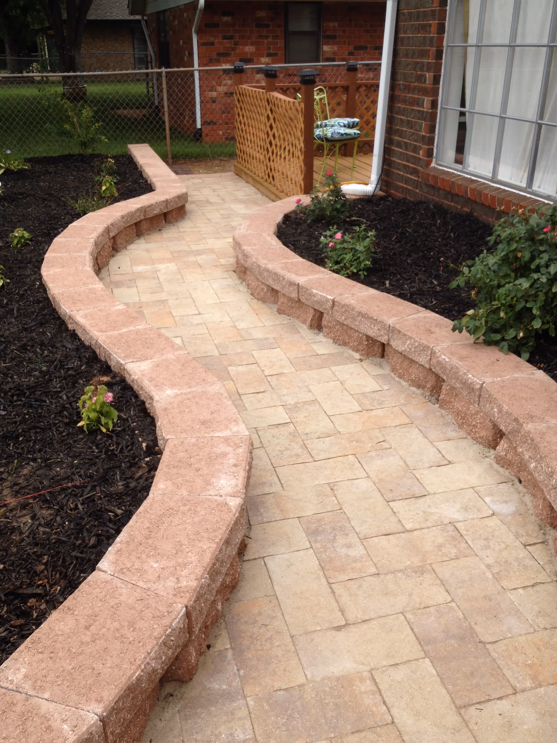 A curved stone pathway bordered by low stone walls with mulch and small plants on either side, leading to a brick house with a window and a small fenced area with a chair and cushions.