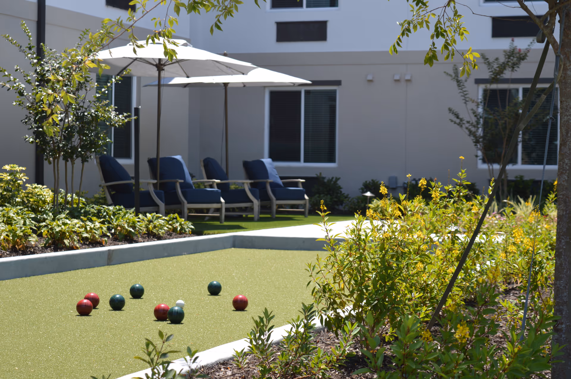Outdoor bocce ball court with several bocce balls scattered on the green surface, surrounded by plants and flowers. In the background, there are three blue cushioned lounge chairs under large white umbrellas, with a beige building and windows behind them.
