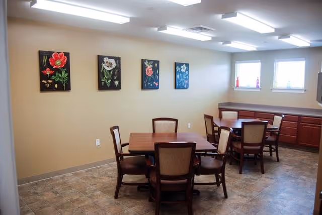 A dining room with two square wooden tables, each surrounded by four cushioned chairs. The walls are beige with four framed floral paintings hanging. There are two windows letting in natural light, and wooden cabinets line one wall beneath the windows. The floor is tiled with a stone pattern.