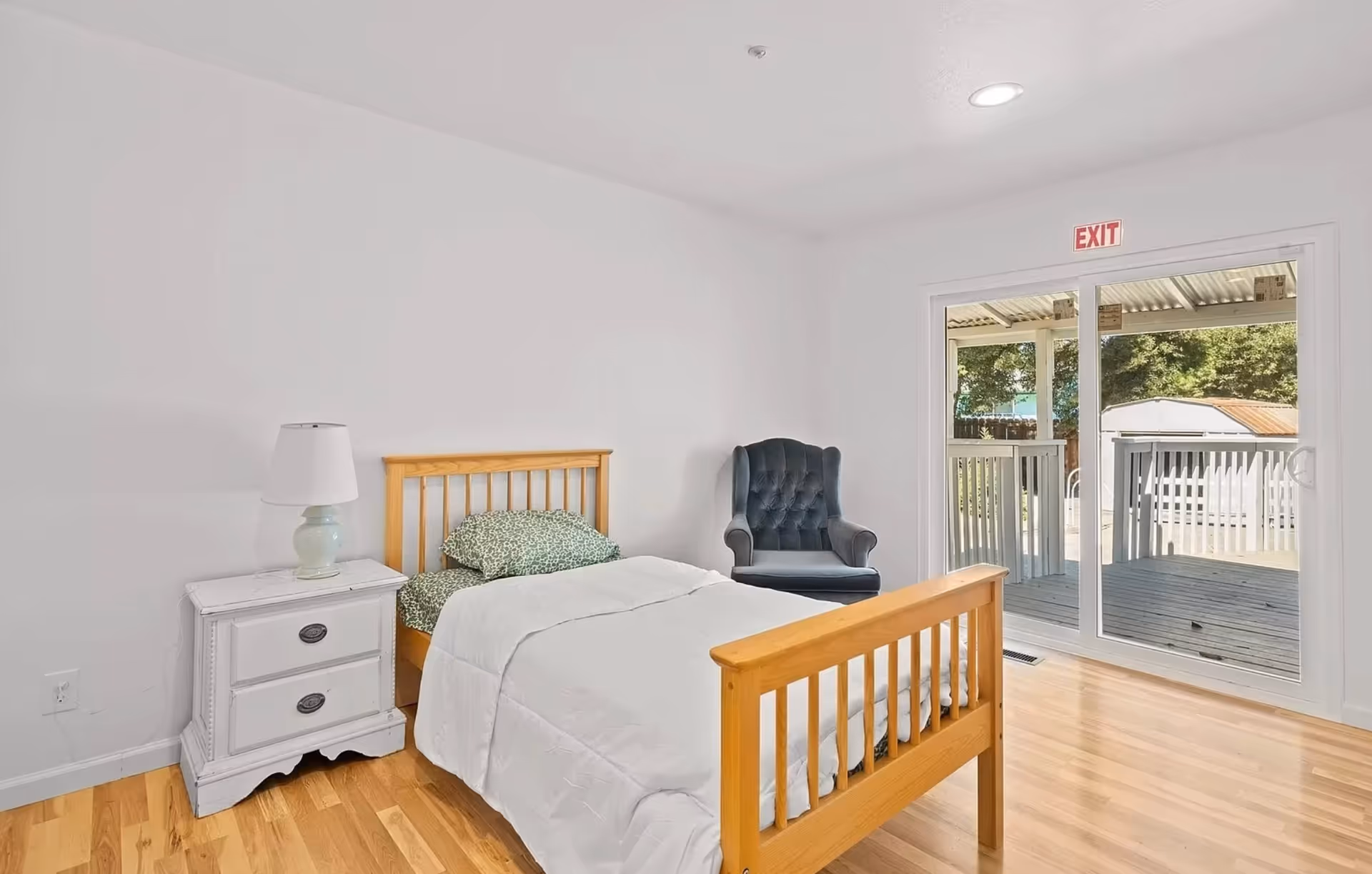 Bright bedroom with wooden bed, nightstand, lamp, chair, and sliding door to a deck.