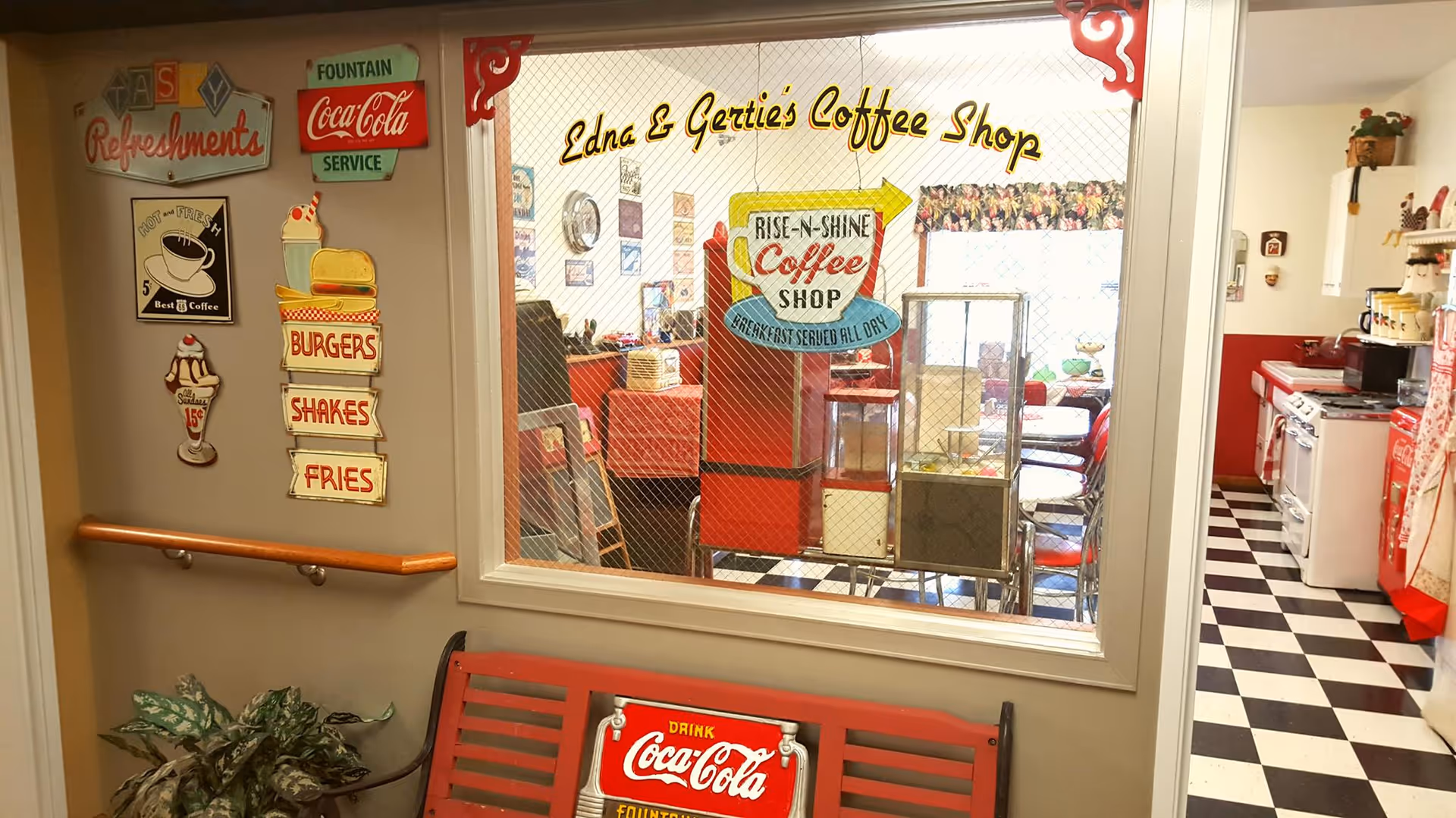 View through a window into Edna & Gertie's Coffee Shop, a retro-style diner with red and white decor, checkered floor, and vintage signs advertising coffee, burgers, shakes, and fries. Outside the window is a red bench with a Coca-Cola sign and a plant on the left side.