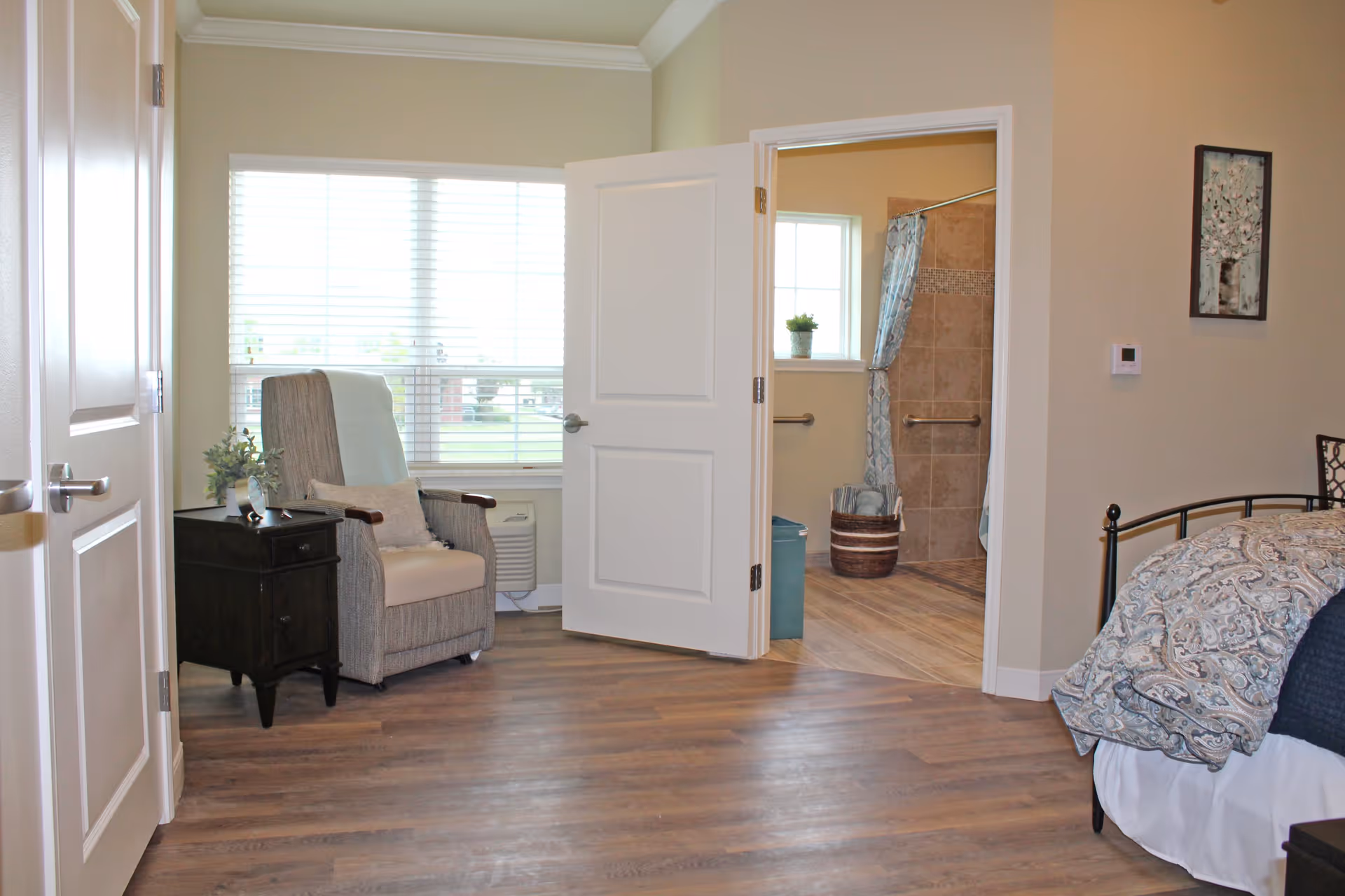 A cozy bedroom with a patterned bedspread on a metal bed frame, a gray upholstered armchair with a pillow next to a black nightstand holding a small plant, and an open door leading to a bathroom with tiled walls, a shower curtain, and grab bars. The room has wooden flooring and a window with white blinds letting in natural light.