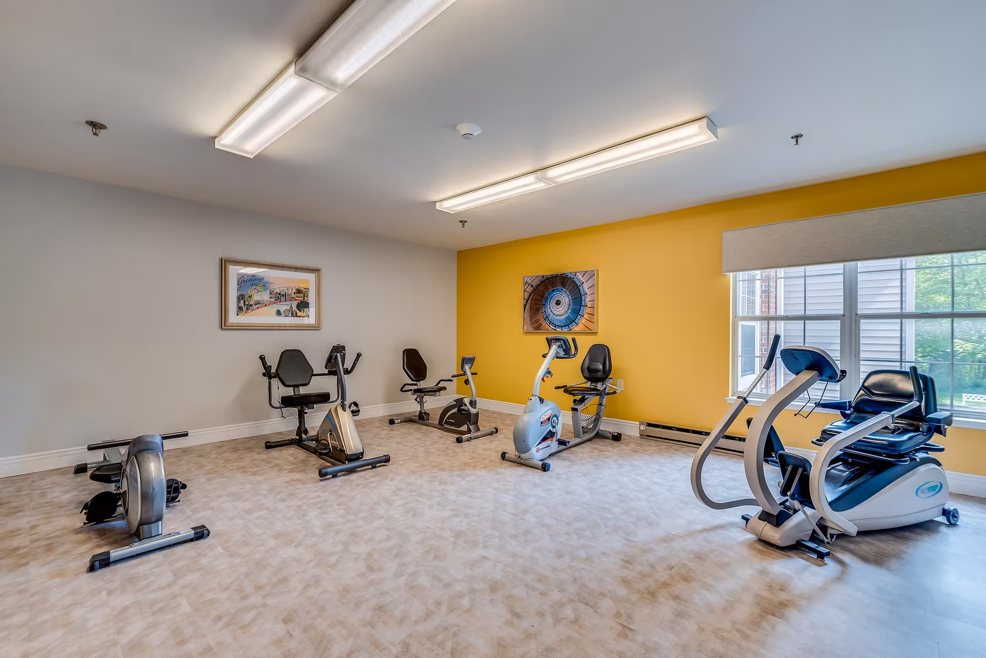 A small exercise room with several pieces of fitness equipment including stationary bikes and a rowing machine. The room has a yellow accent wall with a framed picture and a window letting in natural light. The floor is covered with light-colored patterned vinyl, and the ceiling has fluorescent lighting.