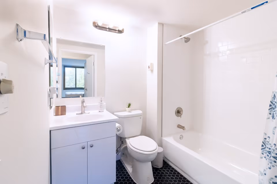 Bright and clean bathroom with a white vanity and sink, a toilet, and a bathtub with a shower curtain. A large mirror is above the sink, reflecting a doorway and a window in the adjacent room.