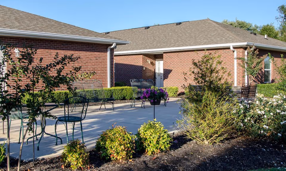 Outdoor patio area with metal chairs and benches on a concrete surface, surrounded by bushes and small trees, adjacent to a red brick building under a clear blue sky.