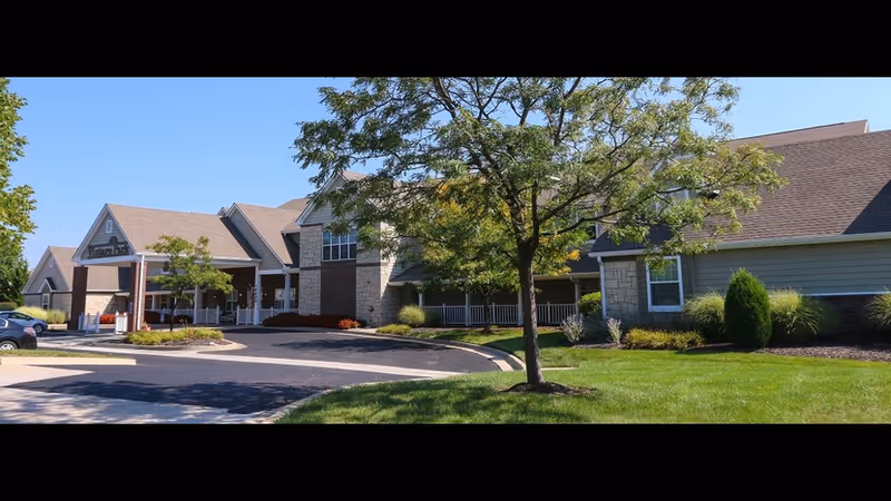 Front exterior of a senior living building with a covered entrance, circular driveway, trees, and a manicured lawn.