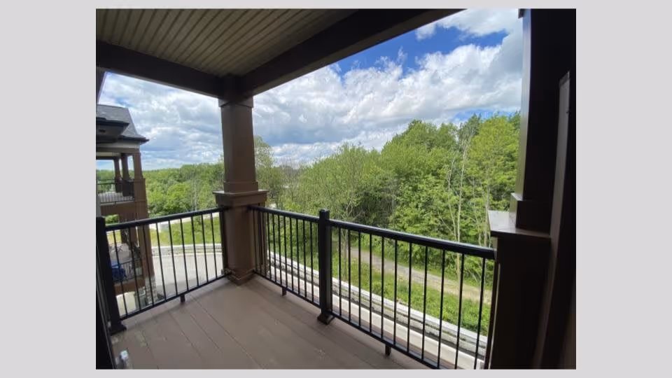 Covered balcony with black metal railing overlooking a green wooded area under a partly cloudy sky.