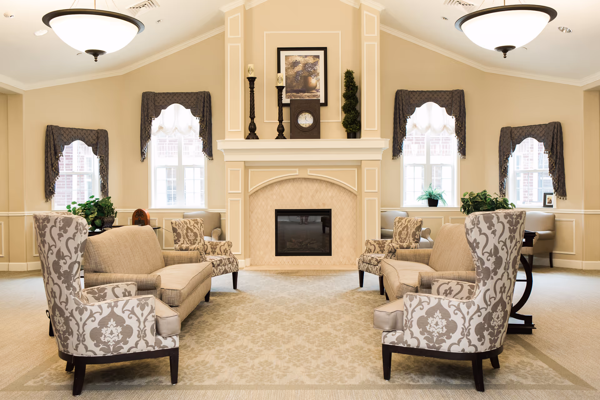 A bright and elegant living room with a beige color scheme featuring two patterned armchairs and two beige sofas arranged around a central fireplace. The fireplace mantel is decorated with candlesticks, a clock, a framed picture, and a small potted plant. Four windows with dark valances allow natural light to fill the room, and there are additional plants on side tables near the windows.