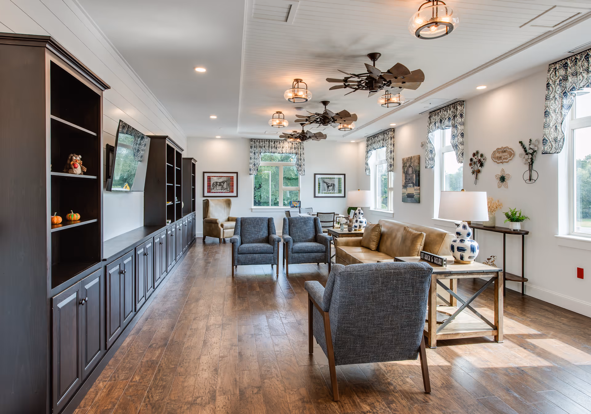 A bright and spacious living room with wooden floors, multiple windows with patterned valances, and ceiling fans with lights. The room features a long dark wood built-in shelving unit with a mounted TV, a tan leather sofa, several gray upholstered armchairs, side tables with decorative lamps, and wall art. Natural light fills the room, creating a welcoming atmosphere.