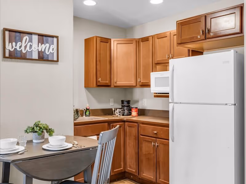 A small kitchen area with wooden cabinets, a white refrigerator, a white microwave, and a coffee maker on the countertop. There is a small dining table set with two place settings and a small green plant. A framed sign on the wall reads 'welcome'.