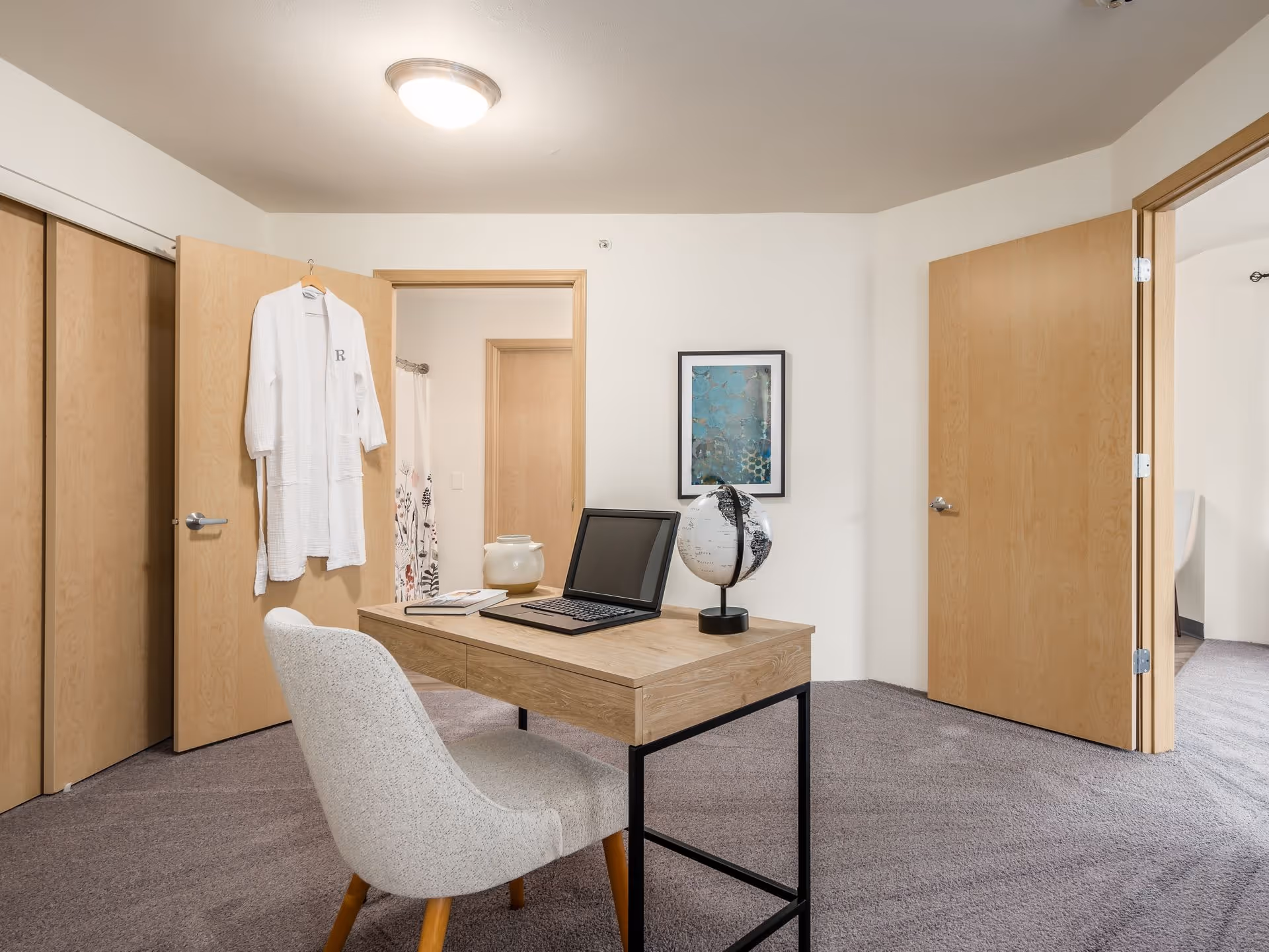Interior room with a wooden desk holding a laptop and globe, a gray upholstered chair, open wooden doors, and a robe hanging on one door.
