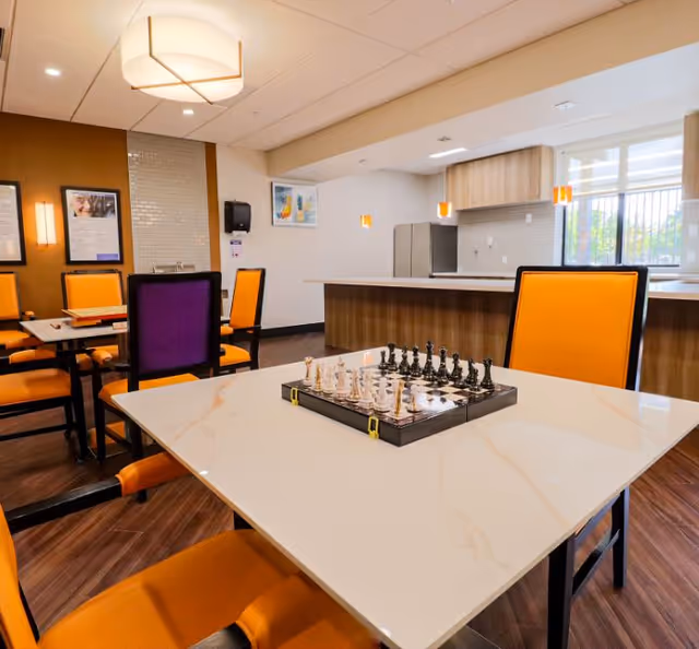 A bright and modern common area with a marble-topped table in the foreground featuring a chess set. The room has orange and purple cushioned chairs, wooden flooring, and a kitchen area with a refrigerator and pendant lights in the background.