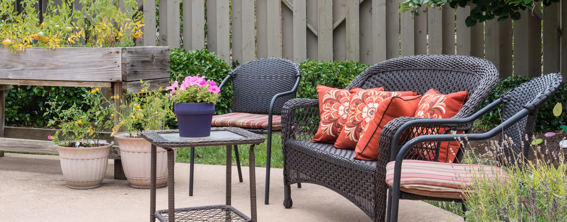 Outdoor patio area with black wicker furniture including a loveseat and two chairs with red and patterned cushions. A small black metal table holds a purple pot with pink flowers. Several potted plants and a wooden raised garden bed are visible against a beige fence in the background.