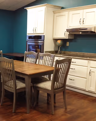 A kitchen area with white cabinets, a built-in oven, teal walls, and a wooden dining table surrounded by chairs.