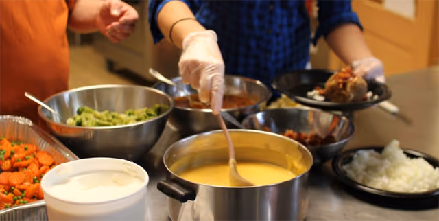 Two people serving food in a kitchen or cafeteria setting. One person is stirring a pot of yellow soup or sauce, while the other is holding a plate with food. Various bowls and trays of food, including cooked carrots, broccoli, rice, and other dishes, are arranged on the counter.