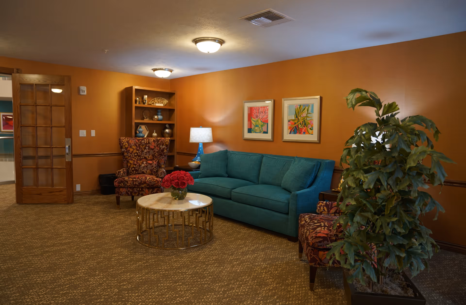 A cozy living room with orange walls, a teal sofa, two patterned armchairs, a round coffee table with a vase of red flowers, a wooden bookshelf with decorative items, two framed colorful paintings on the wall, a table lamp, and a large potted plant.