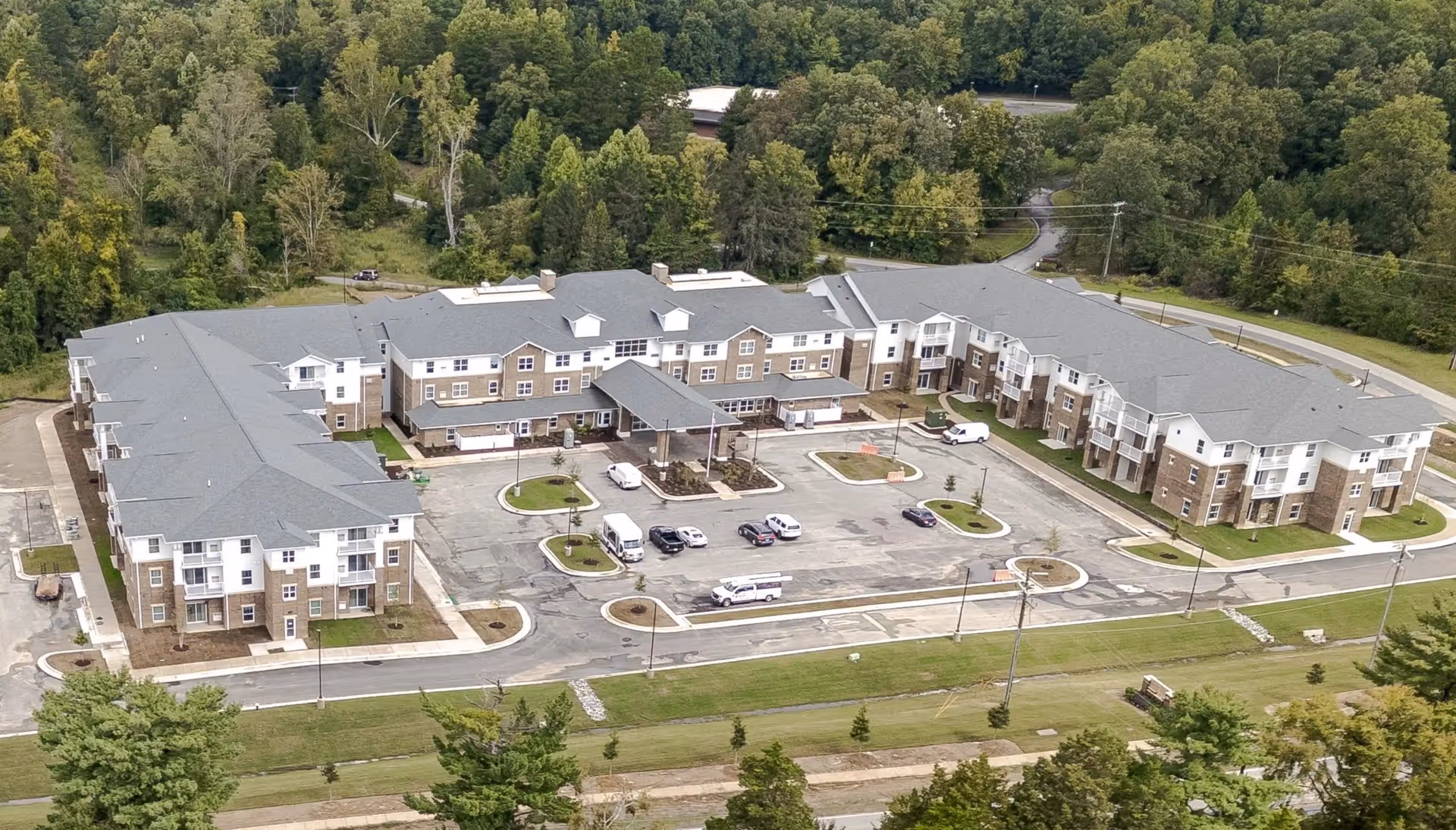 Aerial view of Carolina Pines Retirement facility showing a large, three-story building with a gray roof and brick and white exterior walls. The building is surrounded by a parking lot with several vehicles and is set against a backdrop of dense green trees and a winding road.