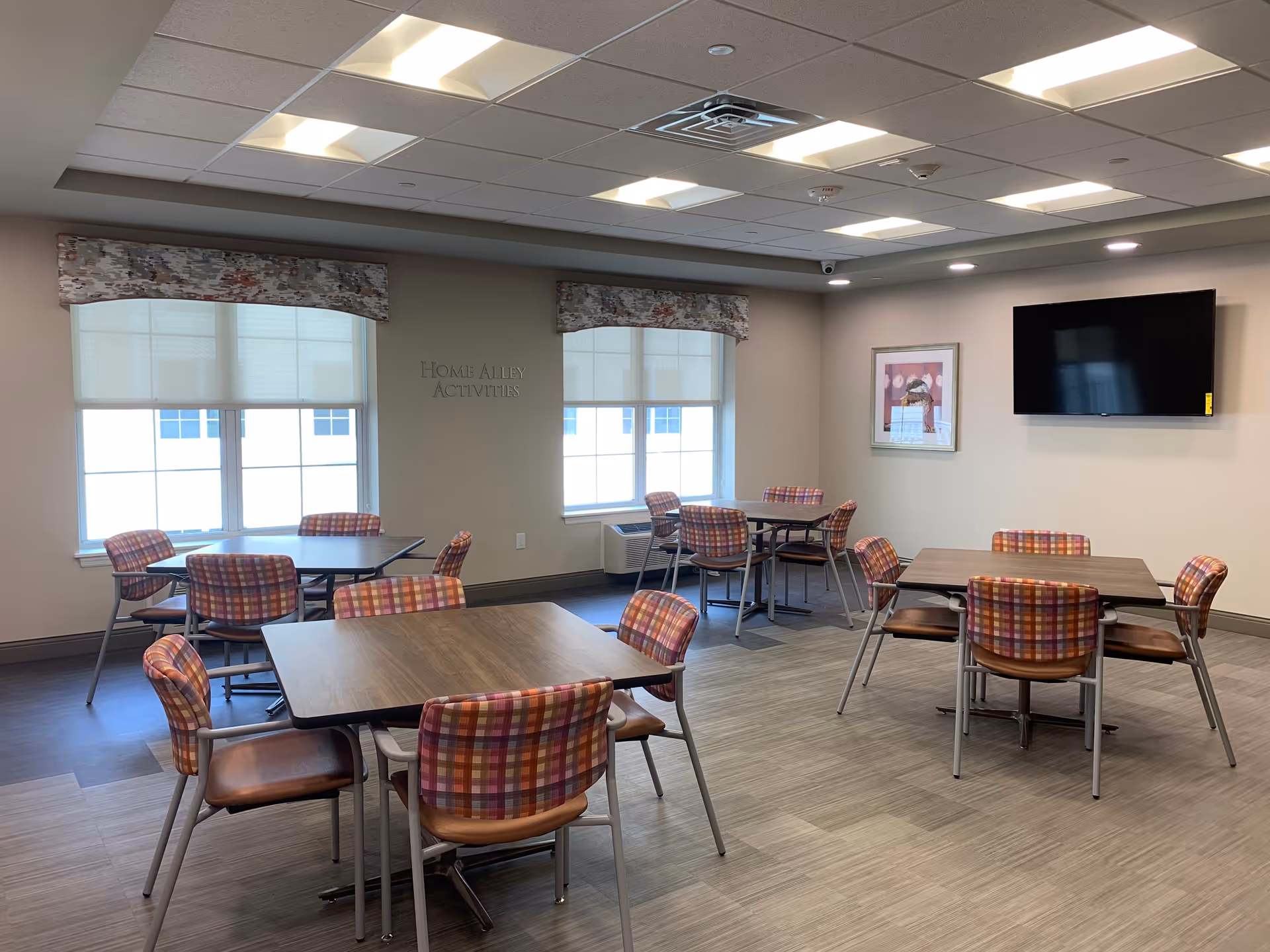 Activity/dining room with several tables and plaid-upholstered chairs, large windows and a wall-mounted TV.