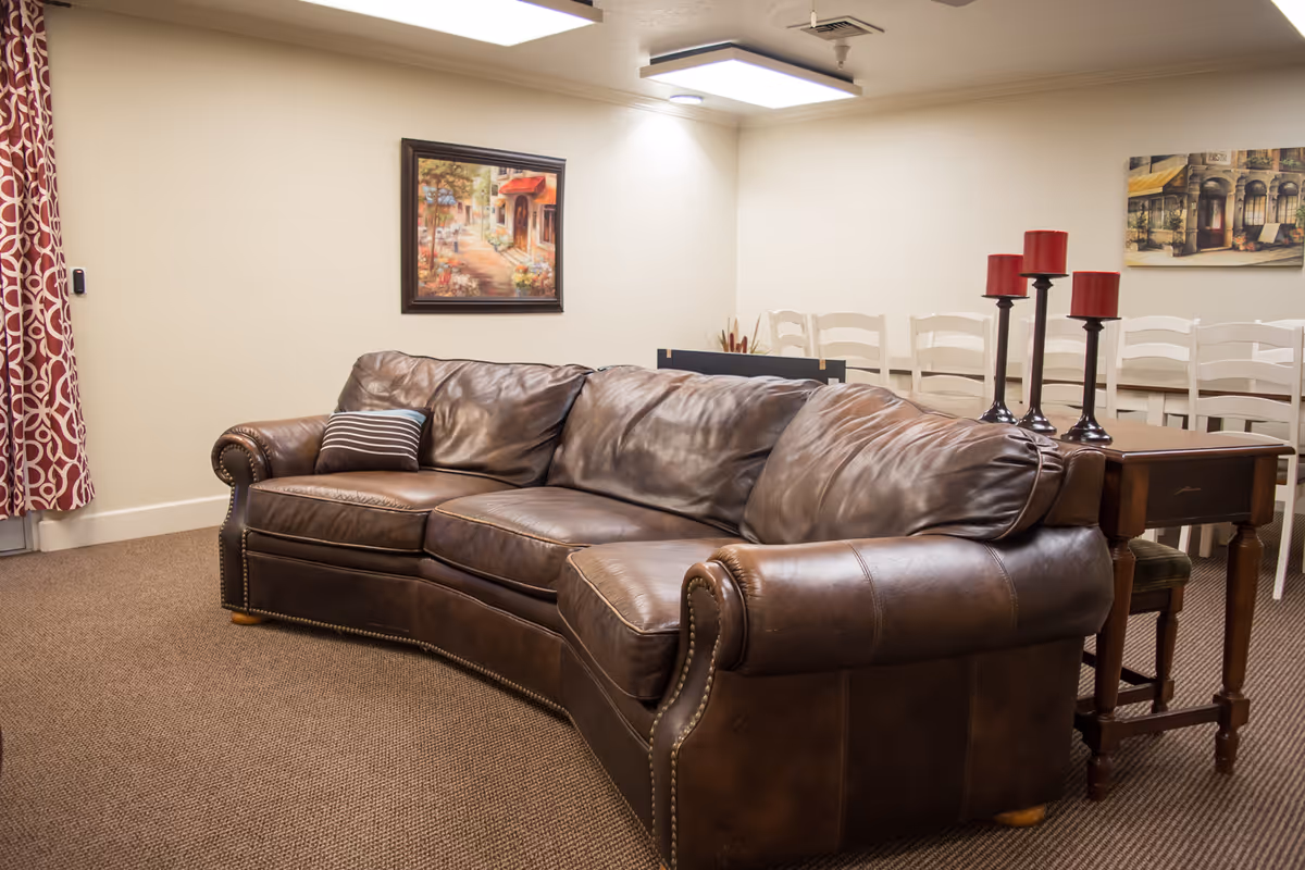 A cozy living room area featuring a large brown leather couch with a striped pillow. Behind the couch is a wooden table with three red candles on tall black candle holders. The background shows a row of white chairs and two framed paintings on the beige walls. The room has carpeted flooring and a patterned curtain on the left side.