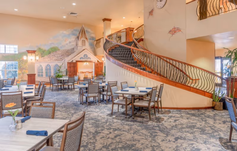 A spacious dining room with multiple tables and chairs arranged neatly. Each table has a small vase with a flower and a folded blue napkin. The room features a large, curved staircase with ornate railings and a wall clock above it. In the background, there is a decorative wall mural depicting a church-like building with open wooden doors and windows, adding a cozy and inviting atmosphere.