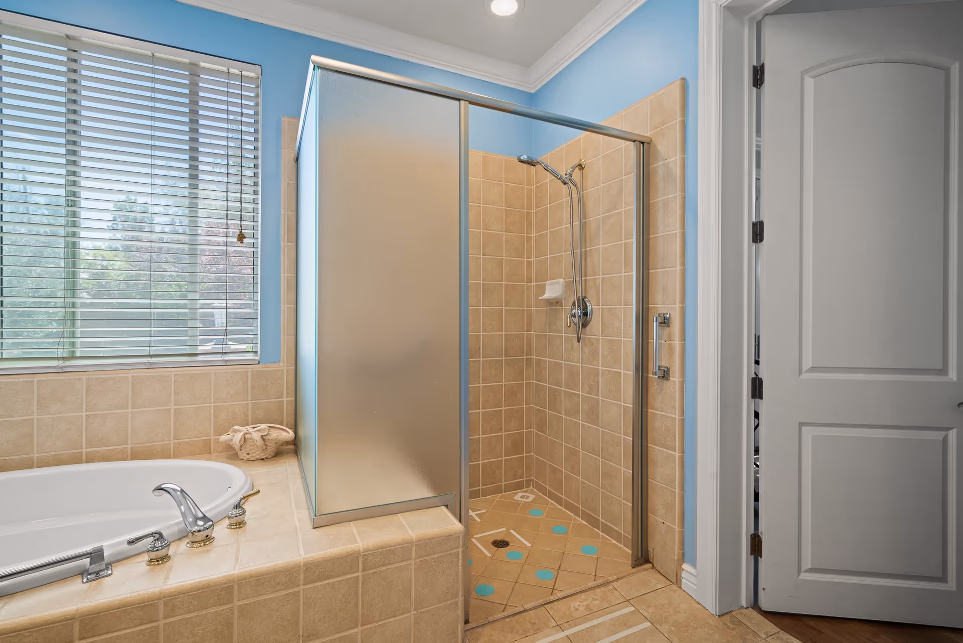 Bathroom with a tiled walk-in shower featuring a frosted glass door and beige tiles. Next to the shower is a large bathtub with chrome fixtures and beige tile surround. A window with white blinds is above the bathtub, and a white door is partially open on the right side of the image. The walls are painted light blue.