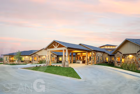 Exterior view of a single-story senior living facility building with stone and wood accents, a covered entrance, and well-maintained landscaping under a colorful sunset sky.