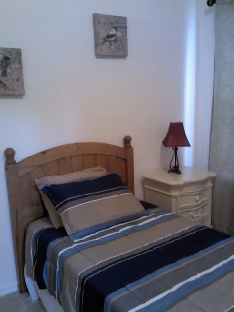 A small bedroom with a single wooden bed featuring a striped blue, gray, and beige bedspread and pillows. Next to the bed is a white nightstand with a red lampshade on top. Two framed bird pictures hang on the white wall above the bed.