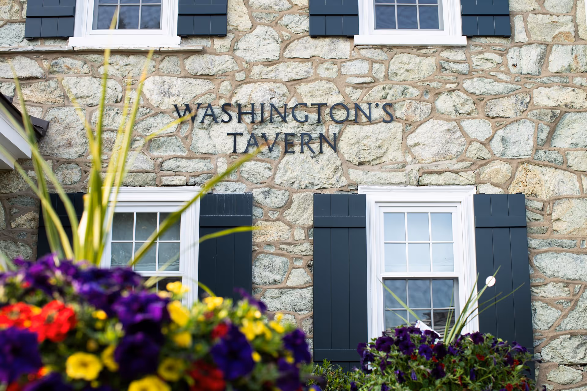 Stone building exterior with two white-framed windows with black shutters. The words 'WASHINGTON'S TAVERN' are mounted on the stone wall between the windows. Colorful flowers and green plants are in the foreground.