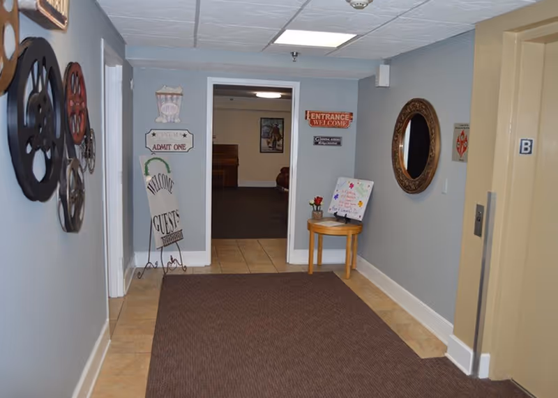 Interior hallway of a senior living facility with light blue walls, a brown carpet runner, and beige tile flooring. On the left wall, there are decorative film reels and a sign that says 'Welcome Guests.' On the right wall, there is a round ornate mirror and a small wooden table with a flower vase and a colorful sign. At the end of the hallway, there is an open doorway leading to another room with a piano and framed artwork on the wall. An elevator door marked with the letter B is visible on the right side.