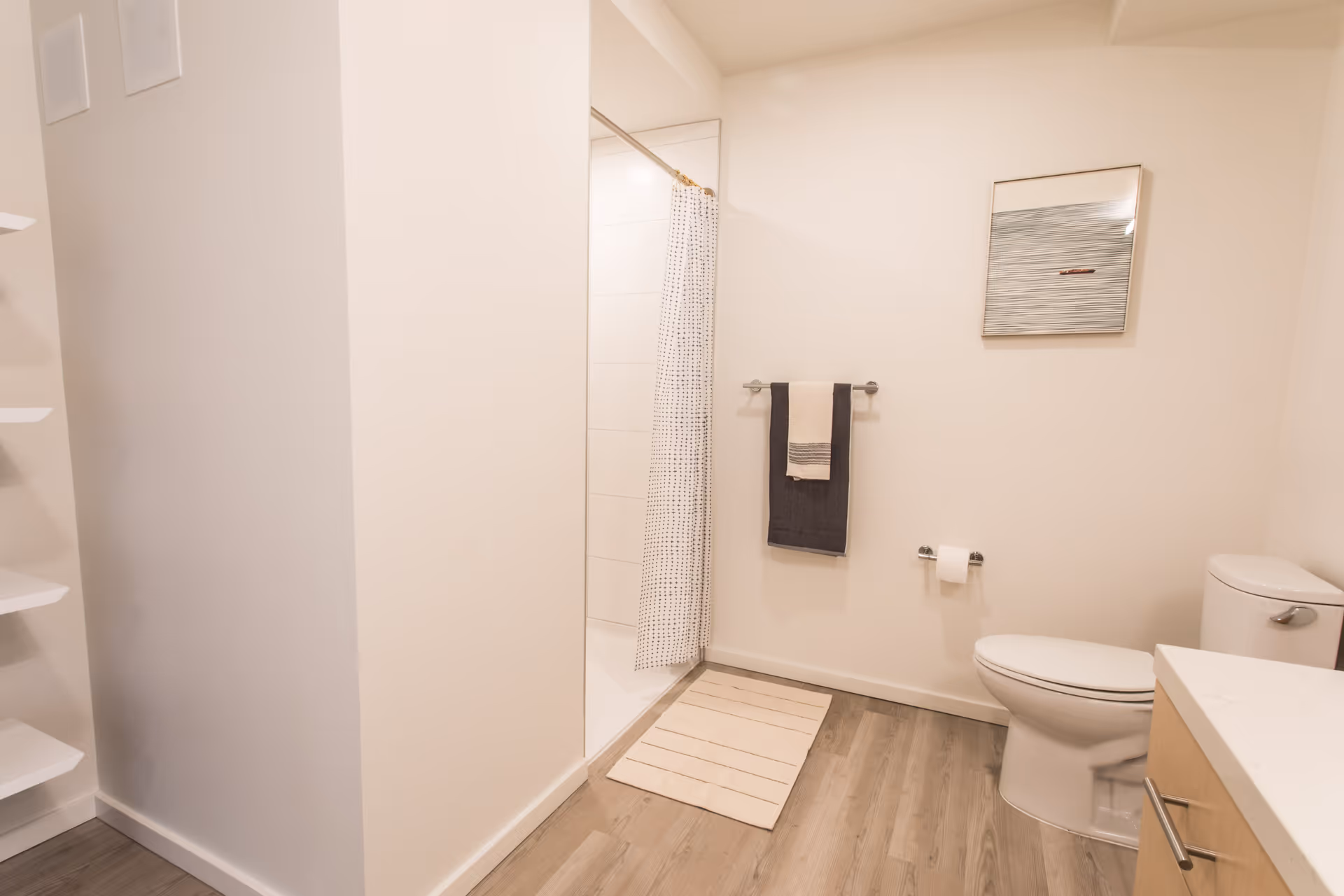 A clean, modern bathroom with light-colored walls and wood-style flooring. The room features a toilet, a shower with a white curtain with black dots, a towel rack with two towels, a small rug in front of the shower, a countertop with a drawer, and a framed abstract artwork on the wall.