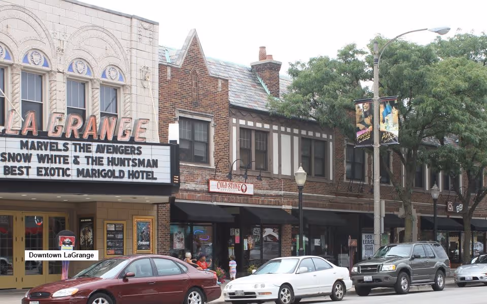 Street view of a downtown area showing a movie theater marquee with the name La Grange and movie titles Marvels The Avengers, Snow White & The Huntsman, and Best Exotic Marigold Hotel. Several cars are parked along the street in front of the theater and adjacent buildings with shops and trees lining the sidewalk.