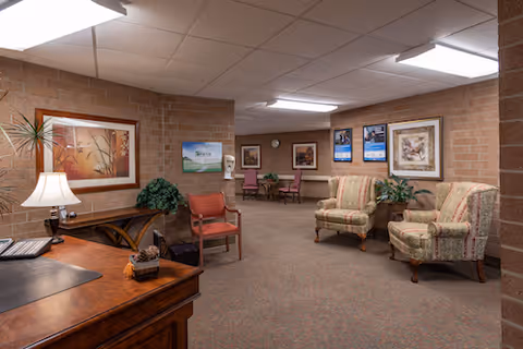 Carpeted reception/lounge area with a wooden desk, upholstered armchairs, framed artwork, and potted plants.