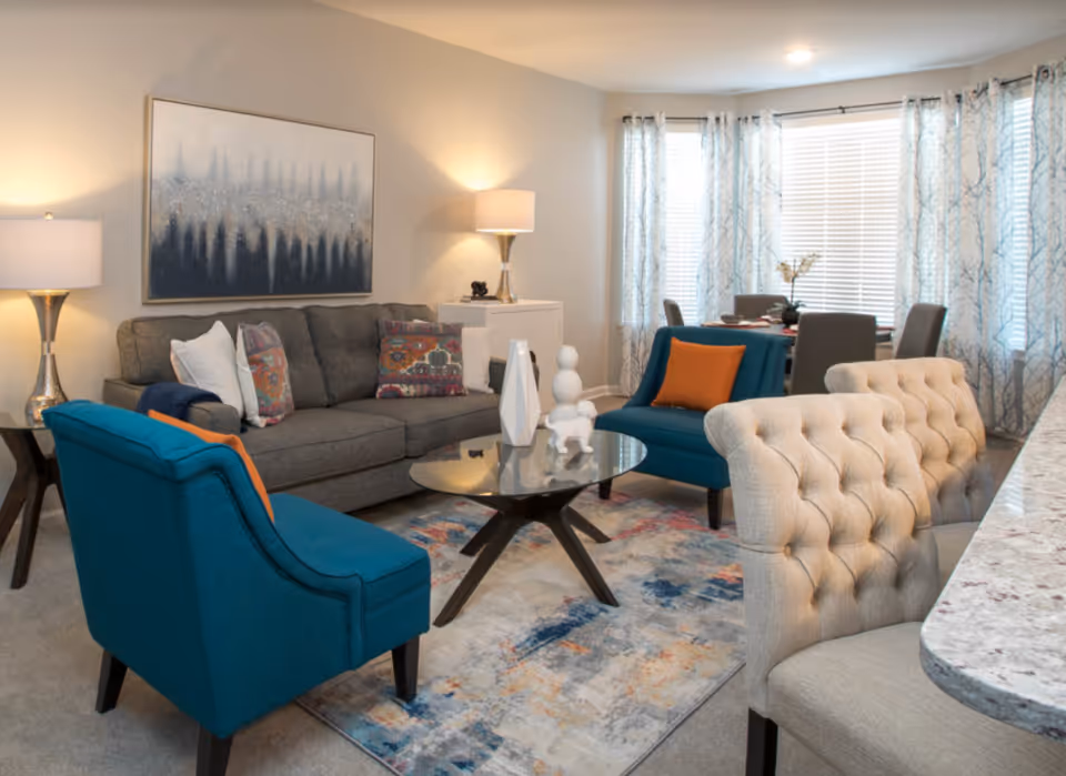 A cozy living room with a gray sofa adorned with colorful pillows, two teal armchairs with orange cushions, a glass coffee table with decorative white sculptures, a patterned area rug, and two beige tufted chairs at a granite countertop. The room is softly lit by two table lamps and natural light coming through large windows with sheer patterned curtains. A large abstract painting hangs on the wall behind the sofa.