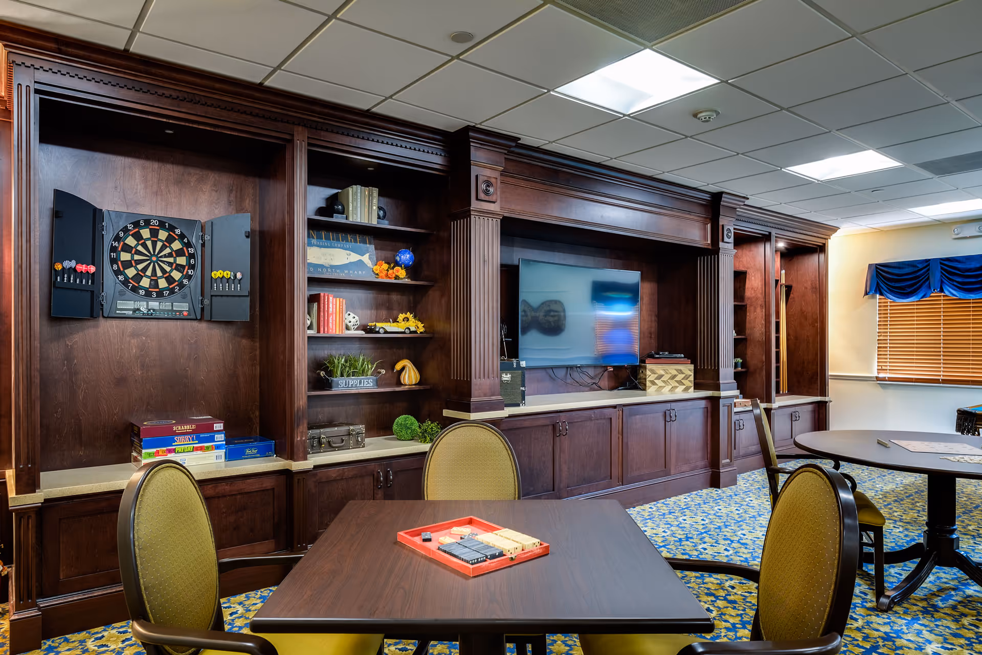 Community recreation room with dark wood built-in shelving and cabinets, a flat-screen TV, dartboard, and tables with chairs.
