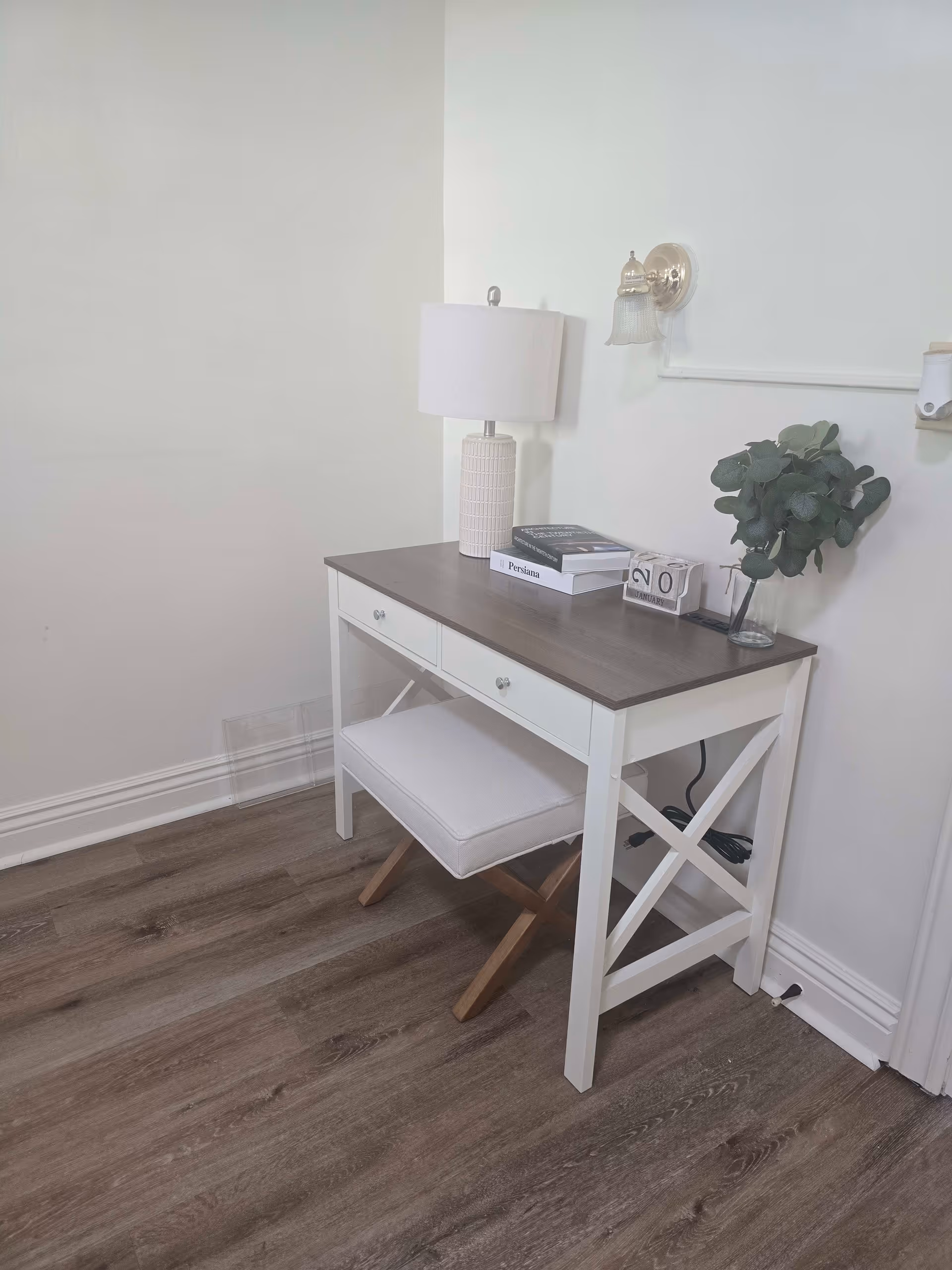 A small white desk with a wooden top against a white wall, featuring a white lamp, two stacked books, a small calendar block showing the number 20, and a glass vase with green foliage. Below the desk is a cushioned stool with wooden legs. The floor is wood with a light brown finish.