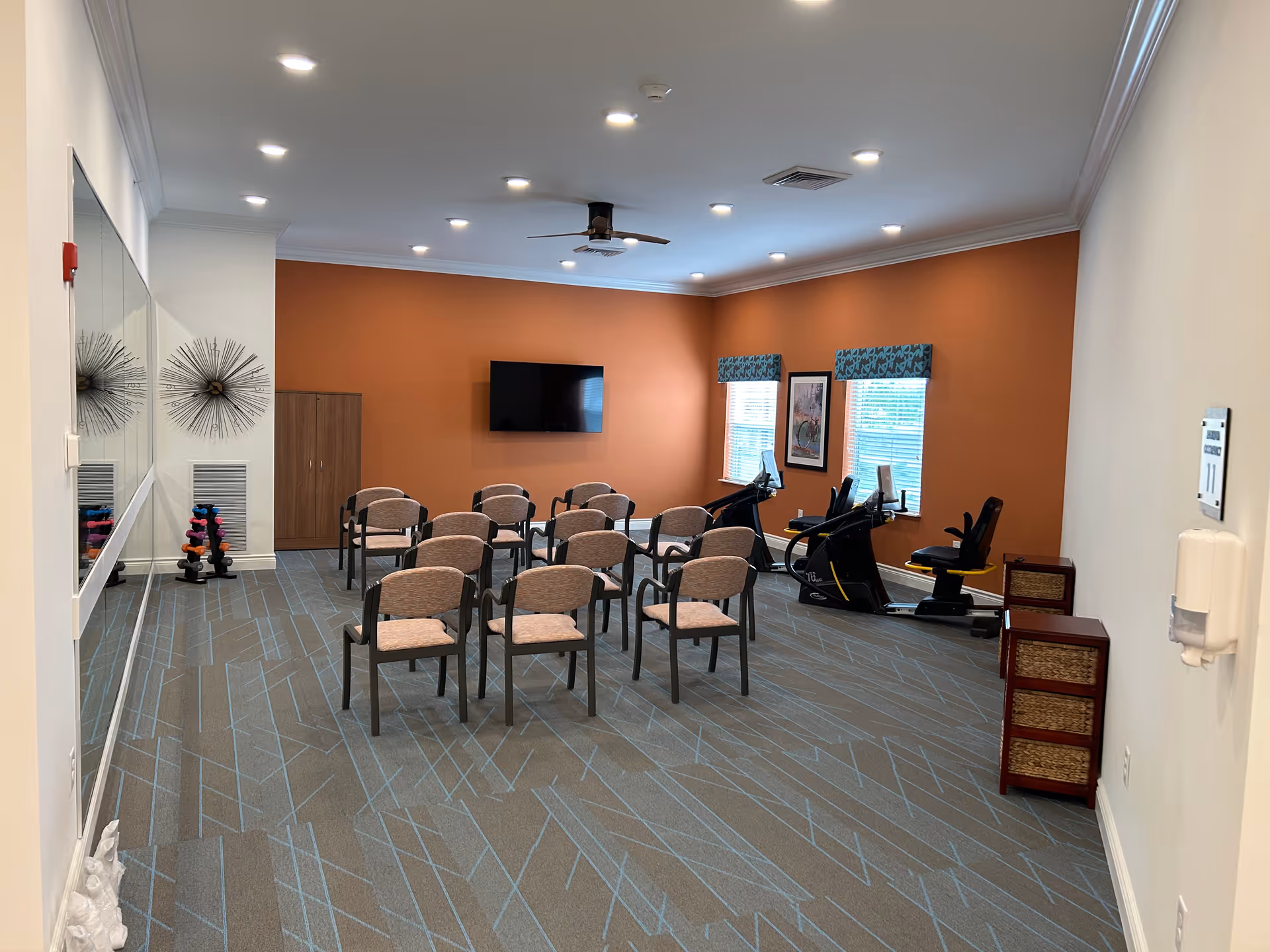 A fitness room with several chairs arranged in rows facing a wall-mounted TV. The room has an orange accent wall, two windows with patterned valances, exercise equipment including recumbent bikes, a rack of dumbbells, and decorative wall art. The floor is carpeted with a geometric pattern.