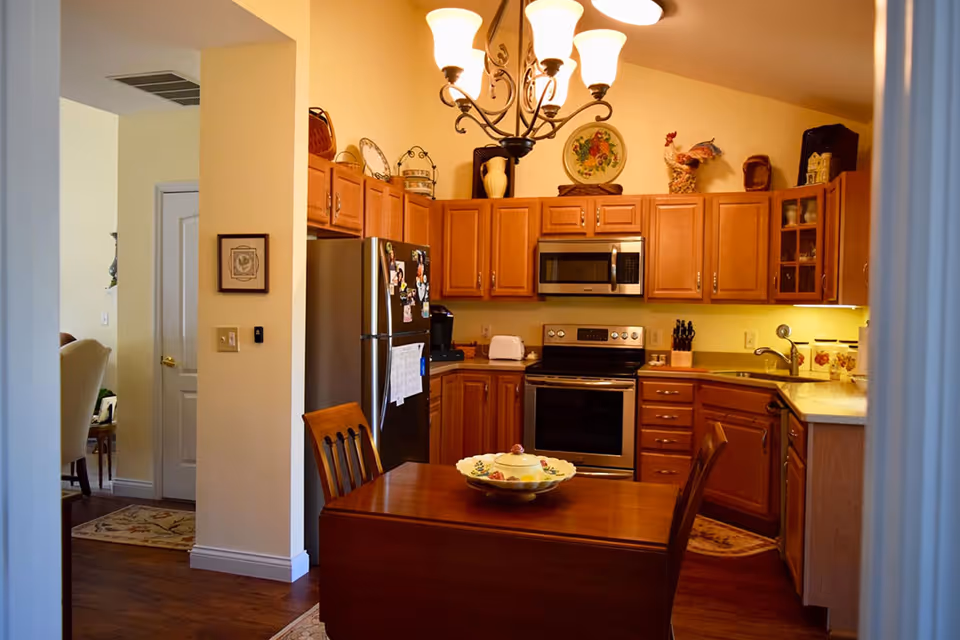 A cozy kitchen with wooden cabinets, stainless steel refrigerator and oven, a microwave above the stove, and a wooden dining table with two chairs. The kitchen is decorated with various items on top of the cabinets and a chandelier hanging from the ceiling. Adjacent to the kitchen is a glimpse of a dining area with upholstered chairs.