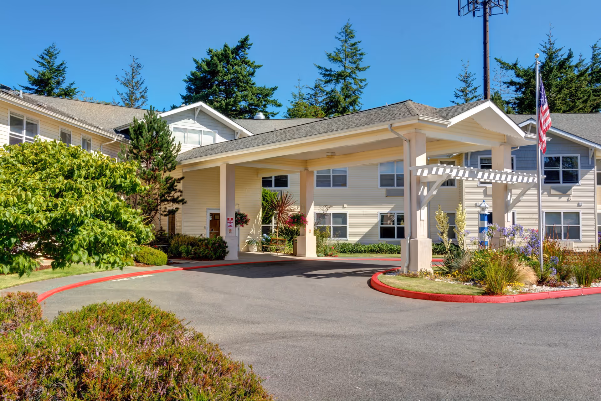 Exterior view of Bayside Terrace by Cogir, showing the entrance with a covered driveway, landscaped greenery, and an American flag on a flagpole under a clear blue sky.