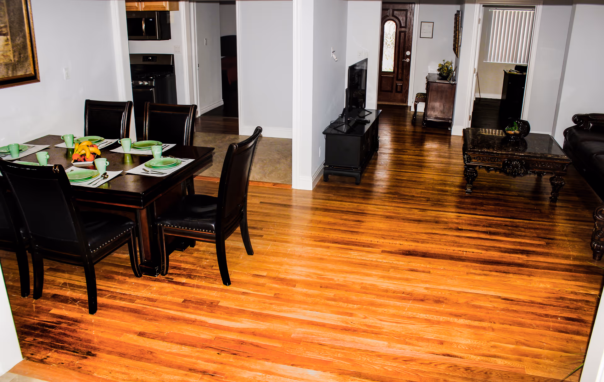 Interior view of a senior living facility showing a dining area with a dark wooden table set for six with green plates and cups, a living room with a dark wooden coffee table and leather sofa, a TV on a stand, and a wooden floor extending throughout the space. The entrance door is visible in the background.