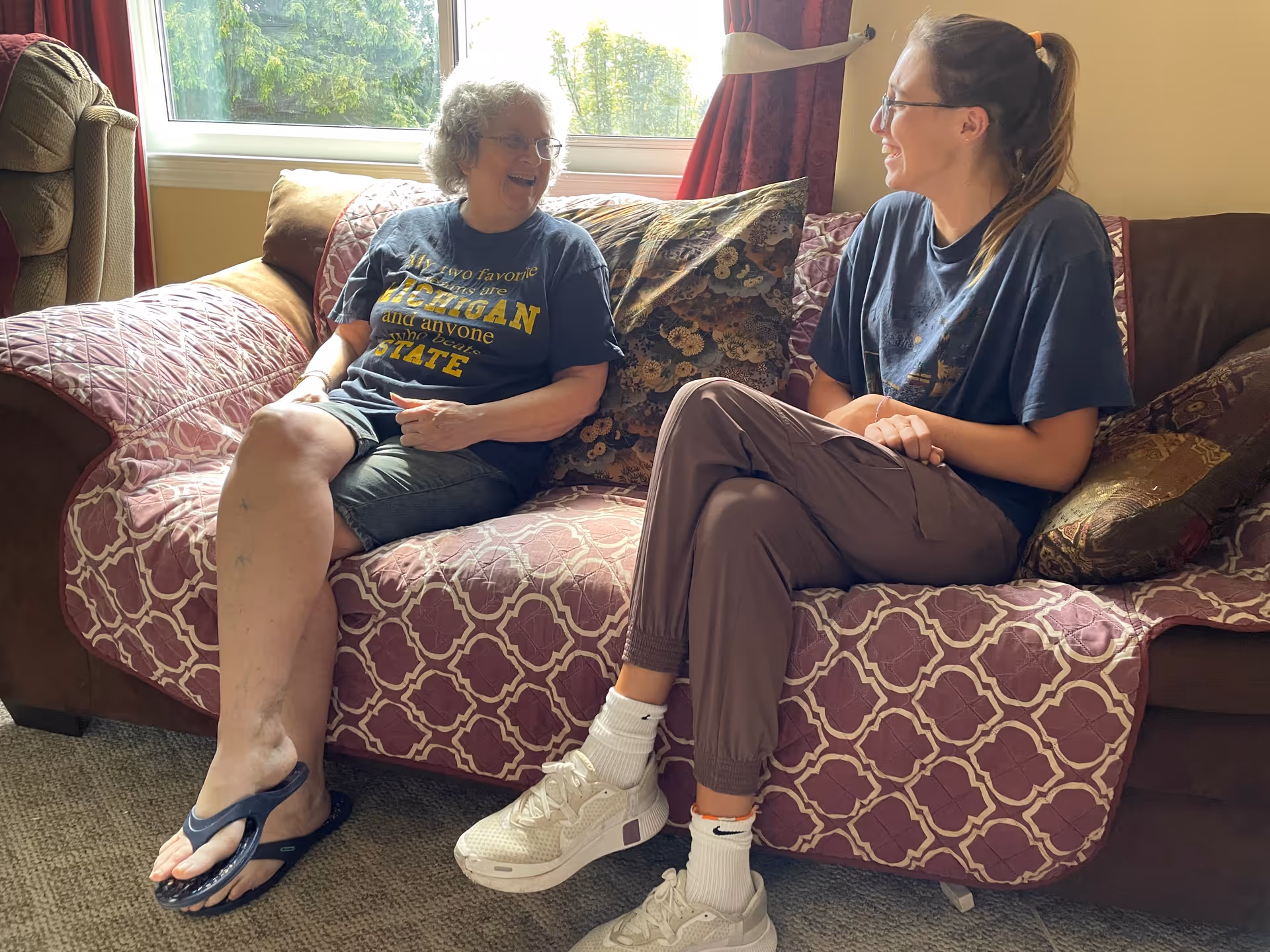 Two people sitting and smiling on a patterned couch in a sunlit living room.